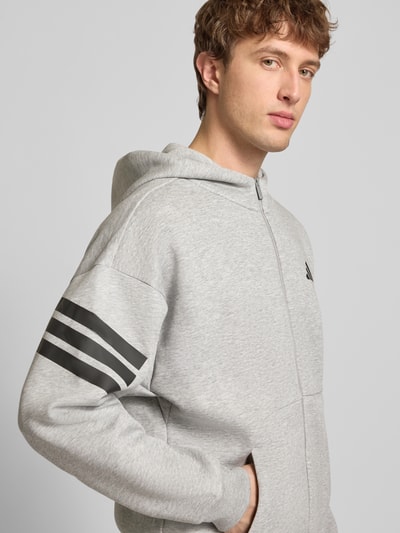 Männermodel trägt Sweatjacke mit Kapuze und Logo von adidas Sportswear, Material-Mix mit Baumwoll-Anteil, Relaxed-Fit, Känguru-Tasche, Logo, Reißverschluss, Farbe Grau, Rückenlänge 68 cm, Ärmellänge 70 cm, Nahaufnahme Oberkörper.
