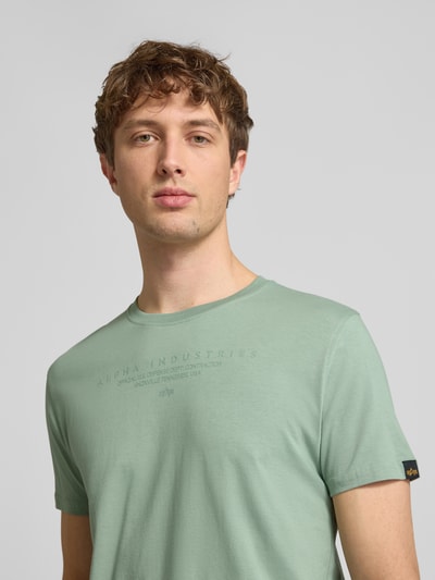 Männermodel trägt T-Shirt mit Label-Print und Rundhalsausschnitt von Alpha Industries in Grün, Reine Baumwolle, Regular-Fit, Nahaufnahme Oberkörper.