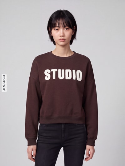 Modelka nosi brązową bluzę z mieszanki bawełny z białym napisem, Loose Fit Sweatshirt aus Baumwoll-Mix Modell 'SWEAT' od ONLY, damski sweter, Ganzkörperansicht von vorne, KI-modifiziertes Bild.