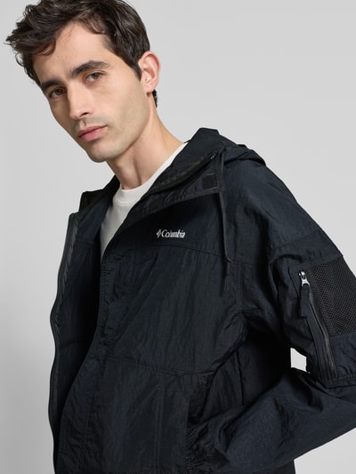 Männermodel trägt Windbreaker mit verdecktem Reißverschluss von Columbia in Schwarz, leicht glänzende Struktur, Logo, Nahaufnahme Oberkörper. Rückenlänge 70 cm, Schulterbreite 64 cm, Brustweite 57 cm, Ärmellänge 56 cm, Regular-Fit, Kapuze mit Tunnelzug.