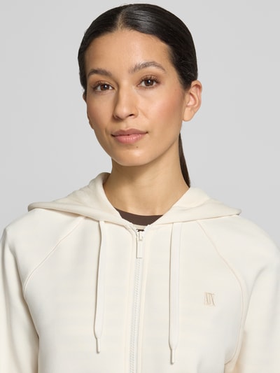 Frauenmodel trägt Regular Fit Sweatjacke aus Viskose-Mix mit Logo-Stitching von ARMANI EXCHANGE in Creme, Nahaufnahme Oberkörper. Elastischer Viskose-Mix, Logo-Stitching, Kapuze mit Tunnelzug, Reißverschluss, elastische Abschlüsse.