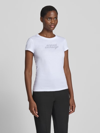 Frauenmodel trägt T-Shirt mit Ziersteinbesatz und Rundhalsausschnitt von Armani Exchange aus reiner Baumwolle, Slim Fit, Logo in Silber auf weißem Stoff, glatte Textur, Damen T-Shirt, Ganzkörperansicht von vorne.
