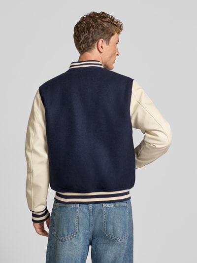 Männermodel trägt College-Jacke aus Woll-Mix mit Eingrifftaschen von Superdry, dunkelblau mit hellen Ärmeln, Rückenansicht. Kontext: Herren College-Jacke von Superdry mit Regular Fit, Stehkragen, Knopfleiste, Rückenlänge 64 cm, Schulterbreite 45 cm, Brustweite 54 cm, Ärmellänge 72 cm.