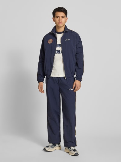 Männermodel trägt Herren Trackjacket mit Label-Stitching Modell 'Slipstream' von PROHIBITED in Dunkelblau, Ganzkörperansicht von vorne, Relaxed Fit, Stehkragen, Reißverschluss, strukturierte Oberfläche, Rückenlänge 64 cm, Ärmellänge 73 cm.