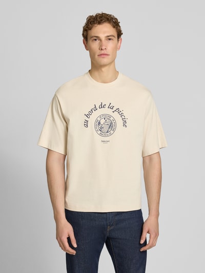 Männermodel trägt Relaxed Fit T-Shirt aus reiner Baumwolle Modell 'BOXYNORMAN GRAPHIC' von SELECTED HOMME in Beige mit Motiv-Print, Ganzkörperansicht von vorne.