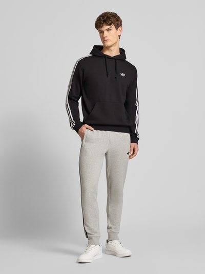 Männermodel trägt Hoodie mit Kapuze von adidas Originals in Schwarz, Baumwoll-Mix, lockerer Schnitt, Streifen, Ganzkörperansicht von vorne.