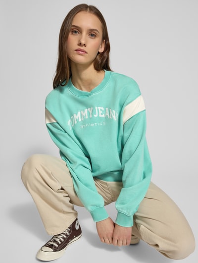 Vrouwenmodel draagt Boxy Fit Sweatshirt uit katoenmix model 'VARSITY CREW' van Tommy Jeans, two tone look met label stitching, geribde contraststrepen en crew neck, kleur turquoise met witte accenten, sweatmateriaal, volledige lichaamsvoorzijde.