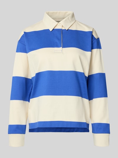 Frauenmodel trägt Regular Fit Langarm-Poloshirt mit verdeckter Knopfleiste von Gant aus reiner Baumwolle, breite blaue und cremefarbene Blockstreifen, Polokragen, Seitenschlitze, Rückenlänge 60 cm bei Größe S, Ganzkörperansicht von vorne.