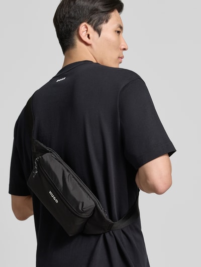 Mężczyzna nosi Bauchtasche mit Label Print Modell 'EVERETT BUMBAG' od HUGO, kolor czarny, suwak, główna komora, nadruk z logo, pasek na biodra z klamrą, całe plecy z tyłu
