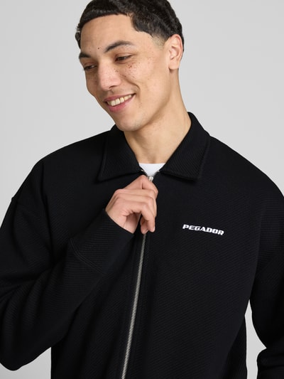 Männermodel trägt Oversized Sweatjacke mit Strukturmuster Modell 'Tate' von Pegador in Schwarz, Baumwoll-Mix mit Stretch-Anteil, Label-Stitching, sichtbarer Reißverschluss, Nahaufnahme Oberkörper.