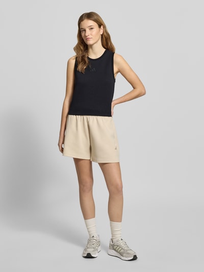 Frauenmodel trägt Regular Fit Top mit Logo-Print von adidas Sportswear in Schwarz, kombiniert mit beigen Shorts, Ganzkörperansicht von vorne. Elastischer Material-Mix mit Modal-Anteil, Rundhalsausschnitt, Rückenlänge bei Größe S: 18 cm.