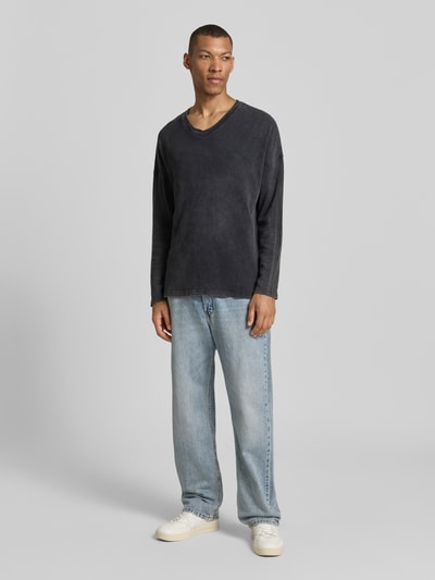 Männermodel trägt Herren Longsleeve mit überschnittenen Schultern Modell 'SONOMA' von American Vintage aus Reine Baumwolle in Regular-Fit, V-Ausschnitt, dunkelgrau, kombiniert mit Jeans und Sneakers, vor grauem Hintergrund, Ganzkörperansicht von vorne. Rückenlänge bei Größe M/L: 66 cm.