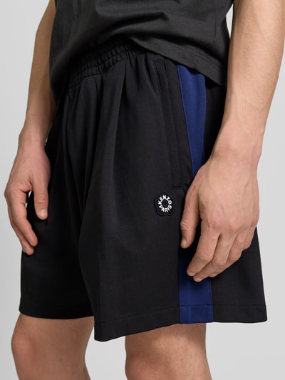 Männermodel trägt Shorts mit Logo-Stitching von Kenzo in Schwarz mit blauen Seiteneinsätzen, elastischer Bund, Eingrifftaschen, Nahaufnahme Oberschenkel. Herren Shorts, Straight Fit, Gesäßtaschen, Innenbeinlänge 46 cm, Bundweite 15 cm.