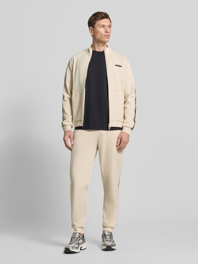 Männermodel trägt Sweatjacke mit Stehkragen und Zweiwege-Reißverschluss von EA7 Emporio Armani in Beige, Regular Fit, Eingrifftaschen, Label-Detail, Rückenlänge 68 cm, Ärmellänge 67 cm, Ganzkörperansicht von vorne.