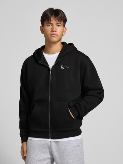Männermodel trägt Sweatjacke mit Label-Stitching und Känguru-Tasche von Karl Kani, schwarz, Baumwoll-Mix, Ganzkörperansicht von vorne.