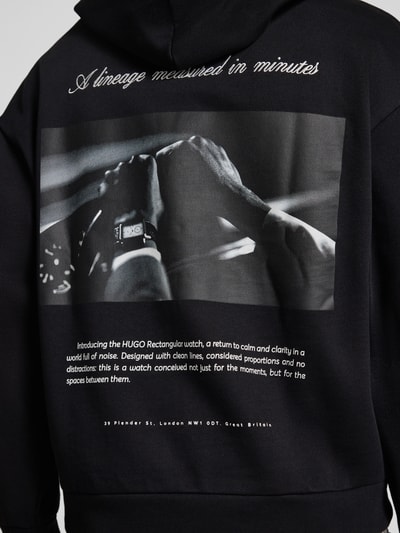 Männermodel trägt schwarzes Oversized Sweatshirt aus reiner Baumwolle Modell 'Devrohood' von HUGO mit Fotoprint auf dem Rücken, Ganzkörperansicht Rückansicht, Herren Sweatshirt von HUGO.