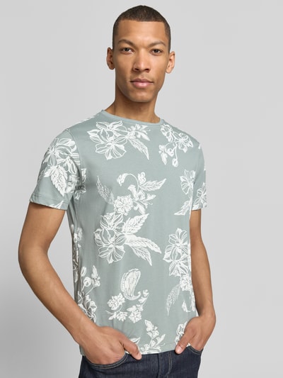Męski model nosi T-Shirt mit Rundhalsausschnitt und Label-Detail od Lerros, bawełna, prosty krój, okrągły dekolt, detal z logo, długość tyłu w rozmiarze M: 70 cm, jasnoszary kolor z białym kwiatowym wzorem, gładka tekstura, całkowity widok z przodu.