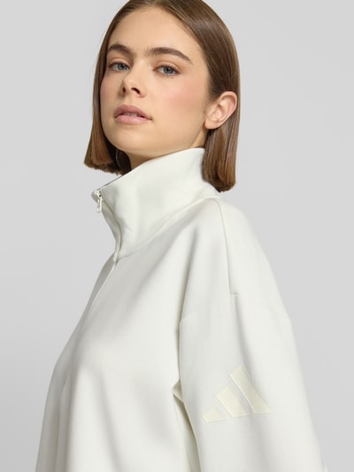 Kobiece model prezentuje Pullover mit Stehkragen und Reißverschluss od adidas Sportswear, biały, logo na rękawie, mieszanka poliestru, ujęcie zbliżeniowe górnej części ciała.
