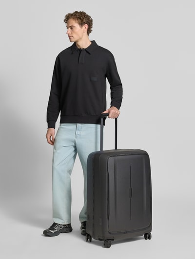 Männermodel trägt Koffer mit Logo-Detail Modell 'Spinner' von Samsonite, Hardshell, schwarz, strukturierte Oberfläche, Ganzkörperansicht von vorne.