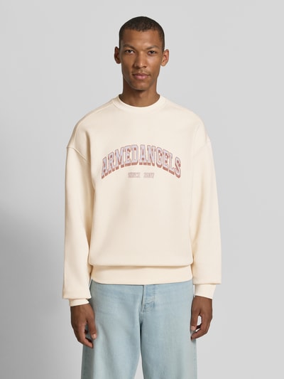 Männermodel trägt Relaxed Fit Sweatshirt aus reiner Baumwolle Modell 'WORDMARK' von ARMEDANGELS in Creme mit rotem Schriftzug, Herren Sweatshirt, Ganzkörperansicht von vorne.