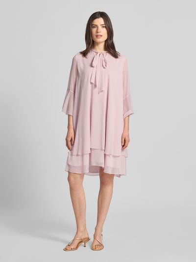 Frauenmodel trägt Knielanges Kleid mit Schleifen-Detail Modell 'Denia' von JOOP!, ausgestellter Schnitt, Lagenlook, zartrosa, fließender Stoff, Stehkragen, Ganzkörperansicht von vorne.