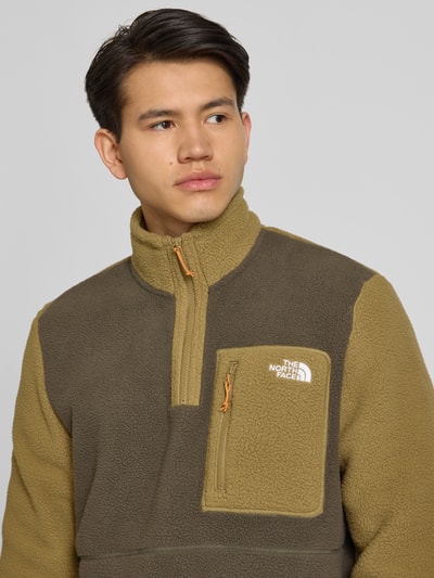Męski model nosi Fleecepullover mit Brusttasche Modell 'YUMIORI' von The North Face, krój regular fit, stójka, wyhaftowane logo, zamek błyskawiczny, brustasza, kolor oliwkowo-beżowy, gładka faktura, długość tyłu w rozmiarze M 70 cm, długość rękawów w rozmiarze M 67 cm, widok z przodu, zbliżenie na górną część ciała.