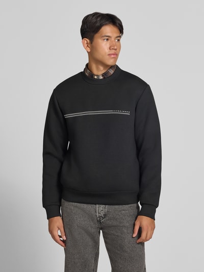 Męski model nosi Relaxed Fit Sweatshirt mit Viskose-Anteil Modell 'Fusion' von JACK & JONES, Mieszanka materiałów z dodatkiem wiskozy, krój relaxed fit, nadruk z logo, krój z długim rękawem, elastyczne wykończenia, długość tyłu w rozmiarze M 66 cm, długość rękawów w rozmiarze M 66 cm, czarny kolor, widok całej sylwetki z przodu.