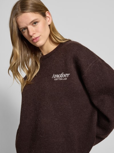 Frauenmodel trägt Oversized Strickpullover mit Rückenmotiv Modell 'Wine & Vibe' von ANOTHER COTTON LAB in Dunkelbraun, weiche Struktur, Rundhalsausschnitt, Label-Stitching, Damen Strickpullover, Nahaufnahme Oberkörper.
