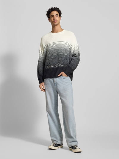 Męski model nosi Oversized Strickpullover mit Woll-Anteil von Another Cotton Lab, mieszanka materiałów z dodatkiem wełny, prążkowane wykończenia, okrągły dekolt, wyhaftowane logo, długość tyłu 68 cm, długość rękawów 55 cm, jasne i ciemne szarości, biel, styl oversize, widok całej sylwetki z przodu