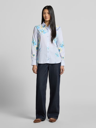 Frauenmodel trägt Regular Fit Hemdbluse aus reiner Baumwolle von Christian Berg Woman mit floralen Allover-Print, hellblaue Grundfarbe, Hemdblusenkragen, langärmeliges Design, kombiniert mit dunkler Hose, Ganzkörperansicht von vorne.