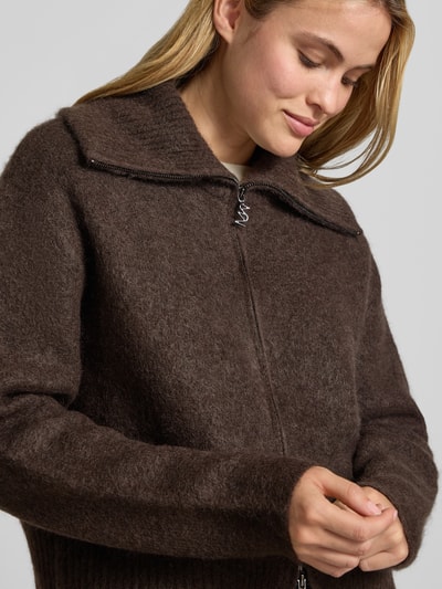 Frauenmodel trägt Strickjacke aus Mohair-Mix mit Label-Detail Modell 'Hulda' von MOS MOSH in Braun, gerippter Bund, Nahaufnahme Oberkörper. Lockere Passform mit Stehkragen, Zweiwege-Reißverschluss, Lanawoll-Anteil sichtbar an feiner, weicher Textur.