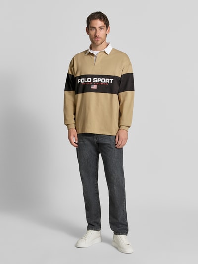 Mężczyzna nosi Relaxed Fit Poloshirt aus reiner Baumwolle od Polo Ralph Lauren, beżowe z czarnym pasem, gładka tekstura, Ganzkörperansicht von vorne.