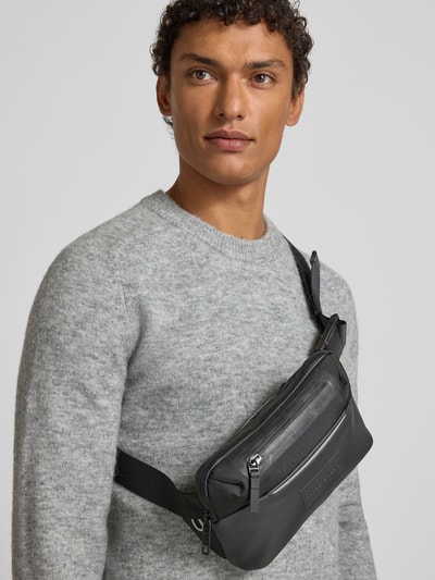Männermodel trägt Tasche mit Label-Detail von Porsche Design in Grau mit reflektierenden Streifen, Reißverschluss und Schulterriemen, Nahaufnahme Oberkörper.