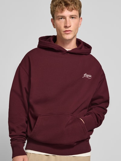 Männermodel trägt Regular Fit Hoodie aus Baumwoll-Mix von PEGADOR in Dunkelrot mit Kängurutasche, Strass-Logo, Nahaufnahme Oberkörper.