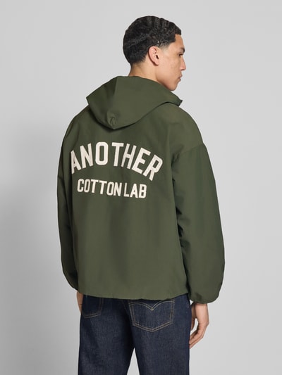 Męski model nosi Adventure Jacke mit Kapuze und verstellbarem Saum od ANOTHER COTTON LAB, zielony kolor, prosty krój, naszywka z logo, dżinsy, widok całej sylwetki z tyłu.