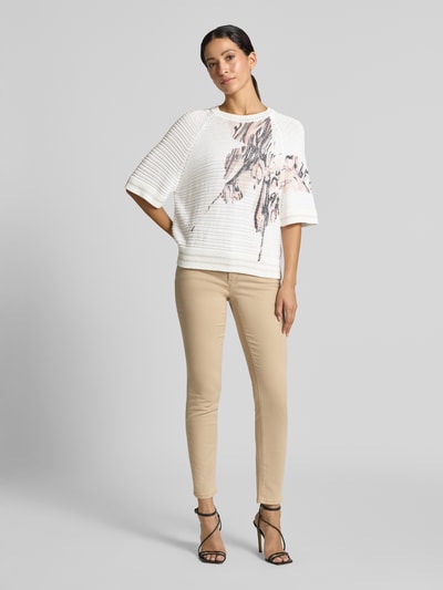 Frauenmodel trägt Strickshirt mit Rundhalsausschnitt von Marc Cain aus Baumwoll-Mix mit Viskose-Anteil im Allover-Look mit Strukturmuster, Regular Fit in Weiß mit beigen und schwarzen Akzenten, Halbarm, kombiniert mit beiger Hose, Ganzkörperansicht von vorne.