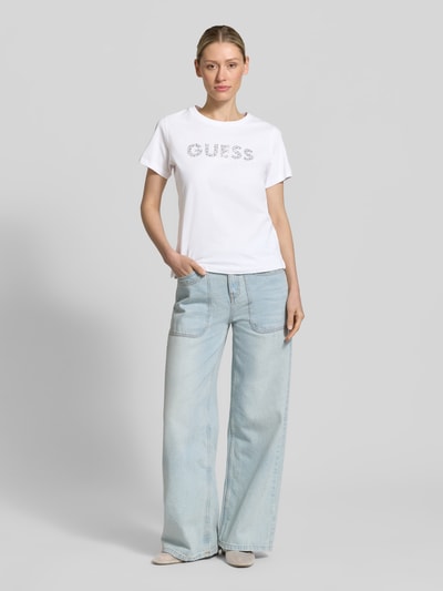 Frauenmodel trägt T-Shirt mit Label-Print und Strasssteinbesatz von Guess aus reiner Baumwolle, Straight Fit, Rundhalsausschnitt, Rückenlänge 59 cm, Ganzkörperansicht von vorne.