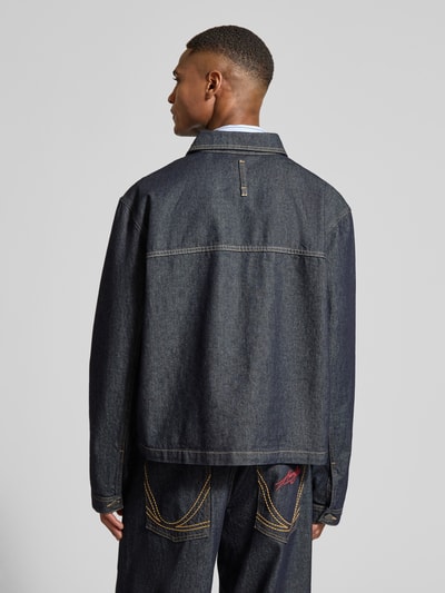 Mannelijk model draagt Relaxed Fit Jeansjacke mit Ziernähten van KARL KANI in donkerblauw met contrastnaden, rugaanzicht, borstzakken, label-stitching, omgeslagen kraag, knoopsluiting, lengte 61 cm, schouderbreedte 50 cm, borstwijdte 65 cm, mouwlengte 69 cm. Ganzkörperansicht Rückansicht.