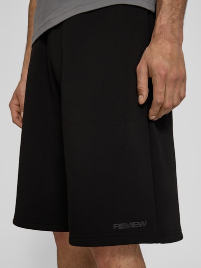 Männermodel trägt Sweatshorts mit Logo-Print von REVIEW in Schwarz, aus Material-Mix mit Baumwoll-Anteil, Loose-Fit, Nahaufnahme Oberschenkel.
