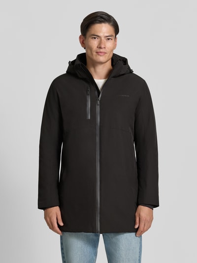 Męski model nosi Regular Fit Parka mit Logo-Print od Lindbergh, elastyczna mieszanka materiałów, czarny kolor, zamek błyskawiczny, kaptur z tunelem, napoleońska kieszeń, długość tyłu w rozmiarze M: 90 cm, szerokość ramion w rozmiarze M: 50 cm, szerokość w rozmiarze M: 58 cm, długość rękawów w rozmiarze M: 65 cm, widok całej sylwetki z przodu.