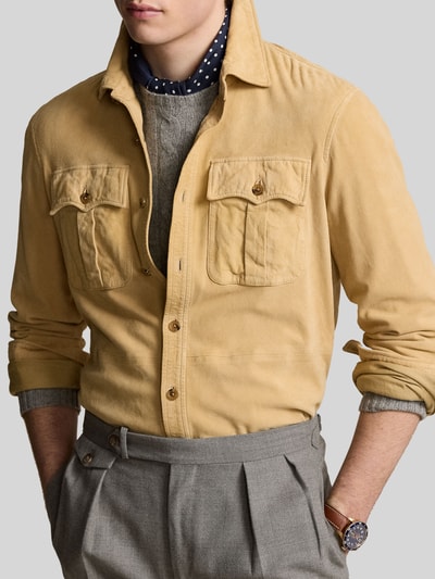 Męski model nosi Suede-Hemdjacke aus Ziegenwildleder od Polo Ralph Lauren, jednolity kolor, kołnierz wykładany, krój regular fit, kieszenie z patką, wykonana z koziej skóry, długość pleców w rozmiarze M 63 cm, szerokość ramion w rozmiarze M 45 cm, szerokość klatki piersiowej w rozmiarze M 41 cm, długość rękawa w rozmiarze M 66 cm, tekstura miękka, kolory beż i szarość, zbliżenie górnej części ciała