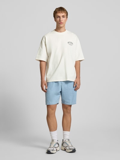 Männermodel trägt T-Shirt mit Label-Detail von REVIEW in Off-White, reine Baumwolle, lockerer Schnitt, Ganzkörperansicht von vorne.