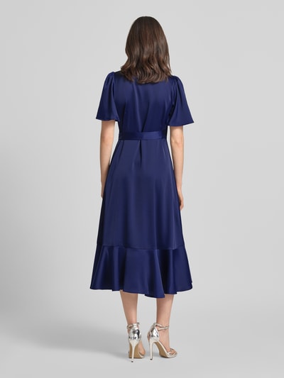 Vrouwenmodel draagt Cocktailkleid mit Volantsaum Modell 'THEA' van YAS in donkerblauw satijn met taillering, midi lengte, volantsaum, vlindermouwen, wikkel look met strik, ruglengte 120 cm, borstbreedte 39 cm, mouwlengte 27 cm, volledige lichaamsweergave achteraan.