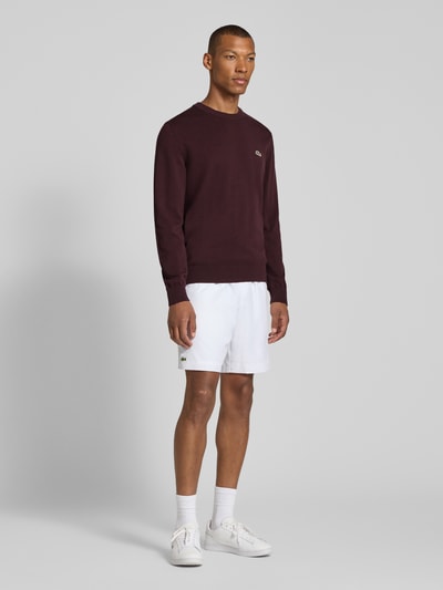 Männermodel trägt Regular Fit Pullover aus Baumwoll-Mix von Lacoste in Dunkelrot und weiße Shorts, Ganzkörperansicht von vorne.