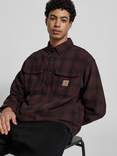 Męski model nosi Regular Fit Freizeithemd mit Logo-Patch Modell 'Edmands' od Carhartt Work In Progress w kolorze bordowo-czarnym, koszula bawełniana z dwiema kieszeniami na piersi, długimi rękawami i kołnierzykiem button-down, szerokość w rozmiarze M 50 cm, długość pleców 76 cm, długość rękawa 61 cm, ujęcie: zbliżenie górnej części ciała z przodu.