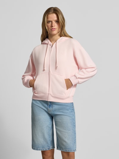 Frauenmodel trägt Relaxed Fit Sweatjacke aus Baumwoll-Mix von Levi's® in zartem Rosa, kombiniert mit blauen Shorts, Ganzkörperansicht von vorne.