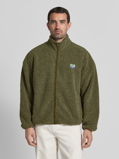 Männermodel trägt groene heren fleecejack met opstaande kraag en labeldetail, relaxed fit, ritssluiting, steekzakken, model 'HOKTOWN' van American Vintage, ruglengte maat M/L 69 cm, schouderbreedte 56 cm, borstwijdte 48 cm, mouwlengte 55 cm, structuur met zachte textuur, geheelopname van voren.