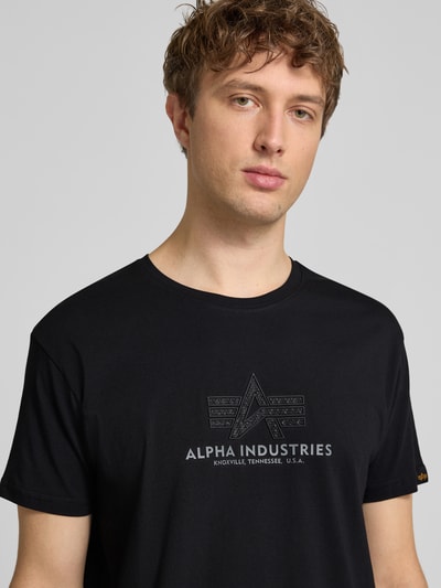Męski model nosi T-Shirt aus Baumwolle Modell 'BASIC' von Alpha Industries, czarny kolor, prosty krój, okrągły dekolt, nadrukowane logo, widok na górną część ciała z przodu.
