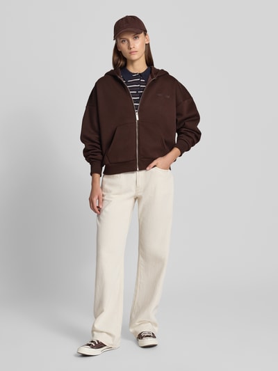 Modelka nosi Relaxed Fit Sweatjacke aus Baumwoll-Mix mit Label-Detail od REVIEW, brązowa bluza dresowa, bawełniany mix, elastyczne ściągacze, podzielona kieszeń kangurka, zamek błyskawiczny, detal z logo, jasne spodnie, przód całej sylwetki.