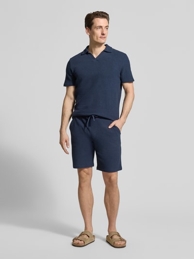 Męski model nosi Regular Fit Poloshirt aus Baumwoll-Mix von MCNEAL w granatowym kolorze, ścieg waflowy, wykładany kołnierz z dekoltem w serek, mieszanka bawełny i elastanu, krótki rękaw, długość tyłu w rozmiarze M: 69 cm, całopostaciowy widok z przodu.